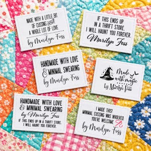 May include: White fabric labels with black text and stitched edges. The labels feature phrases like "Made with love" and "Made with magic" and are signed "by Marilyn Foss". Some labels include a witch hat and bats.