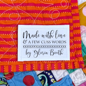 May include: White fabric label with black text that reads "Made with love & a few cuss words by Gloria Booth". The label is sewn onto a colorful striped fabric.