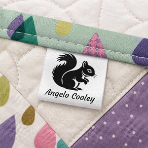 May include: A white label with a black squirrel logo and the text "Angelo Cooley" is sewn onto a quilted fabric. The fabric features a variety of colors and patterns, including green, purple, and pink.