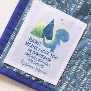 May include: A white label with the text "RAWR MEANS I LOVE YOU IN DINOSAUR" and a blue dinosaur illustration. The label also includes the text "HANDMADE WITH LOVE FOR DUNCAN by Kiana Mercer" and the year "2023". The label is on a blue and white patterned fabric.