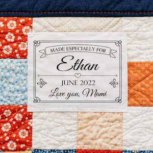 May include: A white label with black text is sewn onto a patchwork quilt. The label reads "MADE ESPECIALLY FOR Ethan JUNE 2022 Love you, Mami" in a decorative font. The quilt features squares of red, blue, and orange fabric.