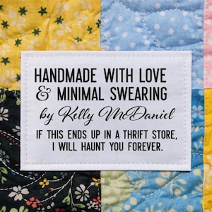May include: A white label with black text that reads "HANDMADE WITH LOVE & MINIMAL SWEARING by Kelly McDaniel. IF THIS ENDS UP IN A THRIFT STORE, I WILL HAUNT YOU FOREVER." The label is on a colorful patchwork quilt.