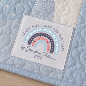 May include: A light blue and white patchwork quilt with a label that reads "MADE ESPECIALLY FOR WILLIAM" above a rainbow design. The label also includes the text "by Grandma Peterson 2022".