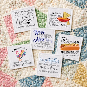 May include: A collection of white fabric squares with various humorous phrases and illustrations printed on them. The squares feature text like "Never Again, This Was a Nightmare!" and "Let's be FRANK". The squares are on a colorful quilt.