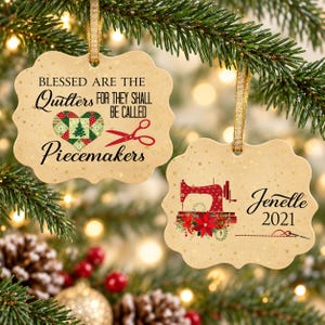 May include: Two scalloped-edge beige Christmas ornaments with gold ribbon hangers. One features a heart-shaped quilt pattern, red scissors, and the text "Blessed are the quilters for they shall be called piecemakers." The other shows a red sewing machine, poinsettias, the name "Jenelle," and "2021."