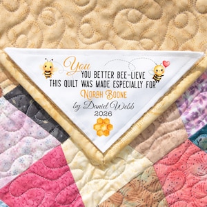 May include: A handmade quilt with a white triangular label. The label features the text "You Better Bee-lieve This Quilt Was Made Especially For Norah Boone by Daniel Webb 2026" with bee illustrations and a honeycomb graphic.