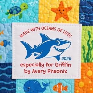 May include: A white fabric label with red and blue text featuring a blue shark illustration. The text reads: MADE WITH OCEANS OF LOVE, 2026, especially for Griffin by Avery Pheonix. The label is stitched onto a colorful quilted fabric with sea life.