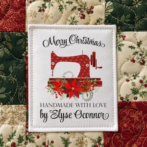 May include: A rectangular white fabric label featuring a red sewing machine illustration with a poinsettia floral arrangement at the base. The text reads Merry Christmas, HANDMADE WITH LOVE, and by Elyse Oconnor in black script and block lettering.