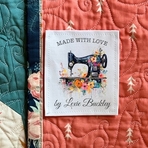 May include: A white fabric label with the text "MADE WITH LOVE" and "by Lexie Buckley". The label features a black sewing machine decorated with colorful flowers. The label is sewn onto a quilt with a floral and geometric pattern.