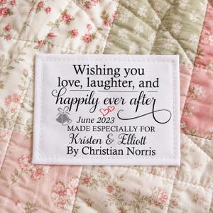 May include: A rectangular white fabric label with black text that reads: Wishing you love, laughter, and happily ever after, June 2023, MADE ESPECIALLY FOR Kristen & Elliott, By Christian Norris, featuring a small bell and heart icon.