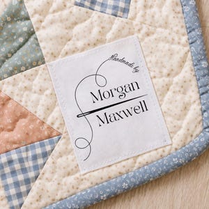 May include: A white fabric label with the text "Handmade by Morgan Maxwell" in black script and a needle and thread design. The label is sewn onto a patchwork quilt with floral and gingham patterns in pastel colors.