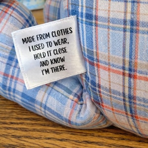 May include: Close-up of a stuffed animal made from blue, white, and orange plaid fabric. A white tag is sewn on the side with the text: "MADE FROM CLOTHES I USED TO WEAR, HOLD IT CLOSE AND KNOW I'M THERE."