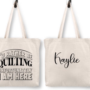 May include: Two white canvas tote bags with black text. One bag says "I'd rather be quilting unfortunately I am here". The other bag says "Kaylie".