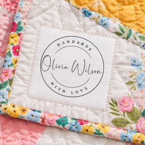 May include: Close-up of a handmade quilt with a floral border and a label that reads "HANDMADE Olivia Wilson WITH LOVE" in a circular design. The quilt features a patchwork design with pink, yellow, and white sections.