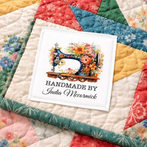 May include: A square white fabric label featuring a blue sewing machine illustration adorned with colorful flowers. The text reads HANDMADE BY India Mccormick in black lettering. The label is stitched onto a multicolored quilted fabric background.