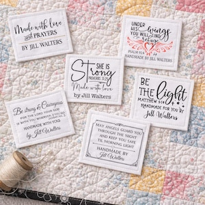 May include: A collection of white fabric squares with inspirational quotes and phrases in black script. The squares are handmade and feature text such as "Made with love and prayers" and "Be the light". A spool of thread and pins are in the image.