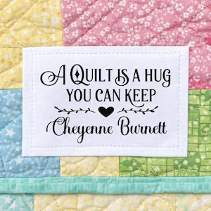 May include: A white rectangular label with the text "A QUILT IS A HUG YOU CAN KEEP Cheyenne Burnett" in black script. The label is on a colorful patchwork quilt with yellow, pink, blue, and green squares.