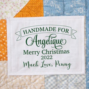 May include: A white fabric label with green text stitched onto a patchwork quilt. The label reads "HANDMADE FOR Angelique Merry Christmas 2022 Much Love, Penny."