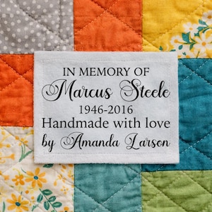 May include: A white fabric label with black text, stitched onto a colorful quilt. The label reads "IN MEMORY OF Marcus Steele 1946-2016 Handmade with love by Amanda Larson". The quilt features squares of orange, yellow, blue, and floral patterns.