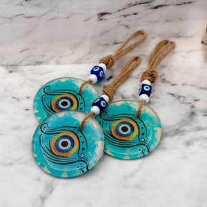 May include: Three round glass ornaments with blue and yellow evil eye designs on a white marble background. Each ornament has a jute string with blue and white evil eye beads.