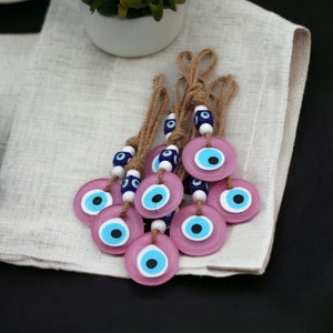 Op de afbeelding: Een groep roze boze oogcharms met blauwe en witte kralen aan een bruin touwtje. De charms zijn op een witte stoffen ondergrond gelegd.