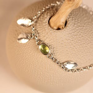 Op de afbeelding: Een zilveren kettingarmband met een enkele ovaalvormige, lichtgroene edelsteen in een gladde zetting, afgewisseld met kleine, platte, getextureerde zilveren blaadjes die aan de schakels van de ketting zijn bevestigd.
