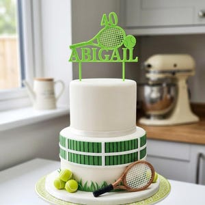 May include: A two-tier white cake decorated with a green tennis-themed topper featuring the number 40, a tennis racket, and the name ABIGAIL. The base tier features a green fence design, three yellow tennis balls, and a miniature tennis racket.