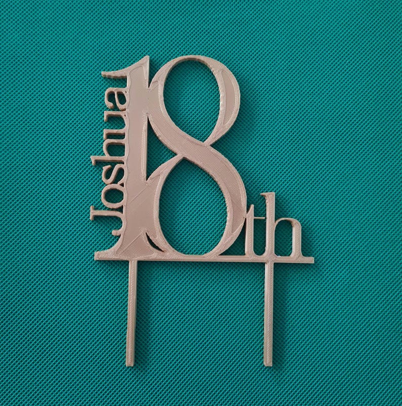 May include: A light pink cake topper with the name "Joshua" and the number "18th". The topper is made of a light pink material and has two support sticks at the bottom. The background is a teal color.