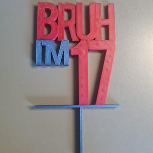 May include: A 3D-printed cake topper in red and blue. The topper reads "BRUH I'M 17" in bold letters. The number 17 is in red, and the other letters are in blue. The topper has a horizontal bar and a vertical stake for insertion into a cake.