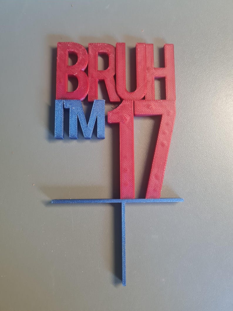 May include: A 3D-printed cake topper in red and blue. The topper reads "BRUH I'M 17" in bold letters. The number 17 is in red, and the other letters are in blue. The topper has a horizontal bar and a vertical stake for insertion into a cake.