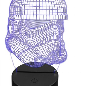 May include: A blue wireframe Stormtrooper helmet night light. The helmet is illuminated with a blue glow and sits atop a black base with a power button. The design is a 3D illusion, creating a unique decorative piece.