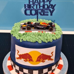 May include: A blue and white cake decorated with a race car, green grass, and a red bull logo. The cake is topped with a blue topper that says "Happy Birthday Corey".