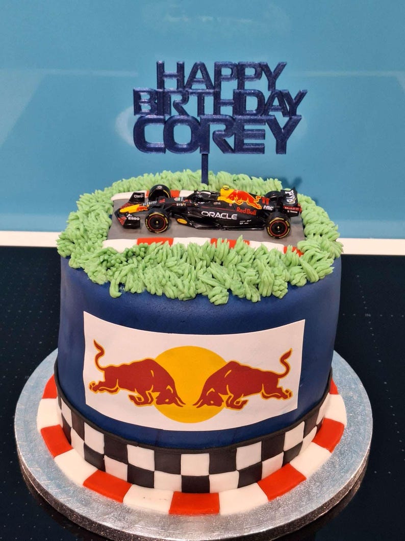 May include: A blue and white cake decorated with a race car, green grass, and a red bull logo. The cake is topped with a blue topper that says "Happy Birthday Corey".