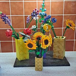 May include: A colorful arrangement of flowers in yellow and gold block-style vases. The flowers include sunflowers, red roses, and other blooms in shades of purple, blue, and yellow. The vases are arranged on a dark surface.
