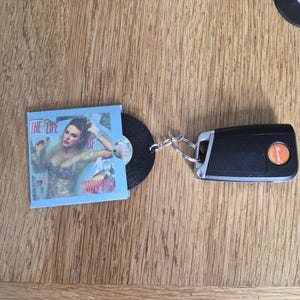 May include: A keychain featuring a black plastic car key fob attached to a miniature vinyl record. The record is partially encased in a light blue paper sleeve with a printed image of a person and the text THE LIFE OF SHOWGIRLS.