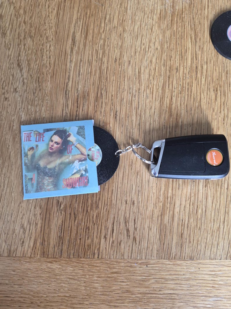 May include: A keychain featuring a black plastic car key fob attached to a miniature vinyl record. The record is partially encased in a light blue paper sleeve with a printed image of a person and the text THE LIFE OF SHOWGIRLS.