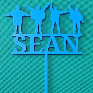 May include: A blue cake topper with four figures in silhouette, arms outstretched, above the name "SEAN". The design evokes a famous band. The topper is made of a solid material and has a stem for insertion into a cake.