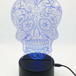 May include: A decorative skull-shaped LED lamp with a blue floral design. The skull is etched with flowers, swirls, and a cross. The lamp sits on a black base that emits a blue glow. A USB cable is plugged into the base.