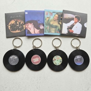 May include: Four square album art prints featuring Band on the Run, The Life, and Thriller, alongside four circular black vinyl record keychains. Each keychain features a central label with distinct imagery, including a mouth with gold teeth.