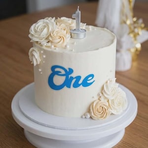 May include: A white frosted cake decorated with buttercream roses and the word "One" in blue script. A silver number one candle sits atop the cake. The cake is on a white cake stand.