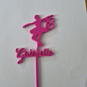 May include: A bright pink cake topper featuring a silhouette of a ballerina in a pose, the number 18, and the name Gabriella in cursive script. The design is attached to a single vertical stake for insertion into a cake or other food item.