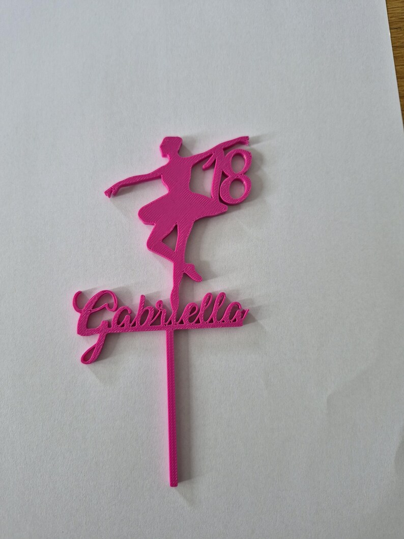 May include: A bright pink cake topper featuring a silhouette of a ballerina in a pose, the number 18, and the name Gabriella in cursive script. The design is attached to a single vertical stake for insertion into a cake or other food item.