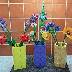 May include: Three vases made of yellow, purple, and gold building blocks hold colorful, artificial flowers. The flowers include red roses, purple lavender, blue and yellow blooms, and a yellow sunflower. The vases are arranged on a dark surface.