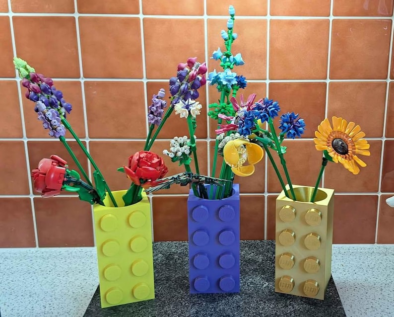 May include: Three vases made of yellow, purple, and gold building blocks hold colorful, artificial flowers. The flowers include red roses, purple lavender, blue and yellow blooms, and a yellow sunflower. The vases are arranged on a dark surface.