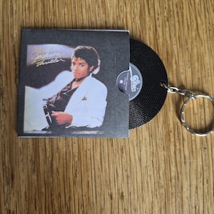May include: A keychain featuring a miniature replica of the Michael Jackson Thriller album cover in a black sleeve, with a small black vinyl record partially emerging from the side, attached to a silver metal split ring for keys.