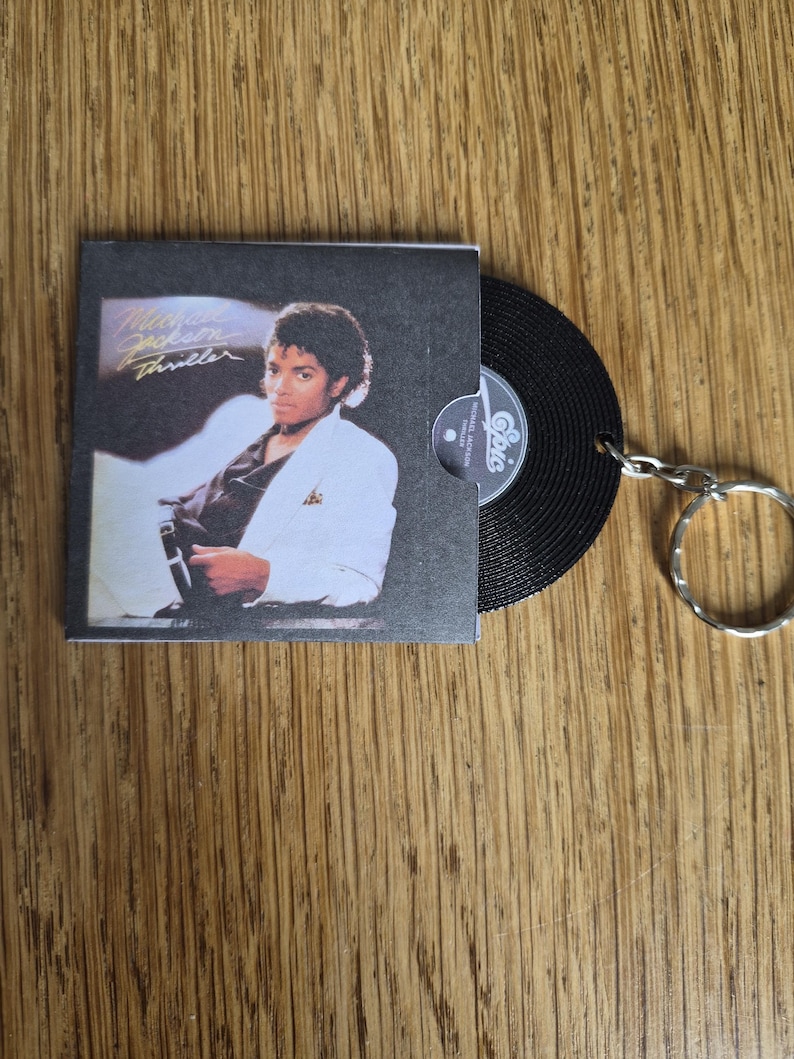 May include: A keychain featuring a miniature replica of the Michael Jackson Thriller album cover in a black sleeve, with a small black vinyl record partially emerging from the side, attached to a silver metal split ring for keys.