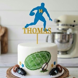 May include: A white frosted cake decorated with a green football field graphic, two small black cleats, and two brown footballs. A blue silhouette of a football player with the number 14 and the name THOMAS in gold sits on top.