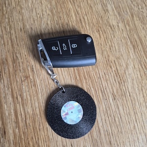 May include: A black car key fob with three buttons attached to a circular keychain shaped like a black vinyl record. The center label of the record features a small graphic with the text The Life of a Showgirl printed in a light-colored font.