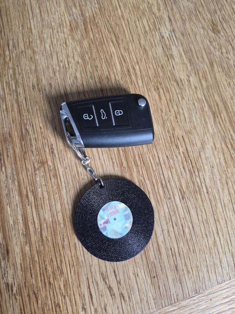 May include: A black car key fob with three buttons attached to a circular keychain shaped like a black vinyl record. The center label of the record features a small graphic with the text The Life of a Showgirl printed in a light-colored font.