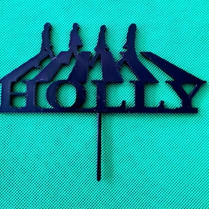 May include: A dark blue cake topper with the name "HOLLY" cut out in a stylized font. The topper features a silhouette of a circus tent with three figures on top. The topper is on a green mesh background.